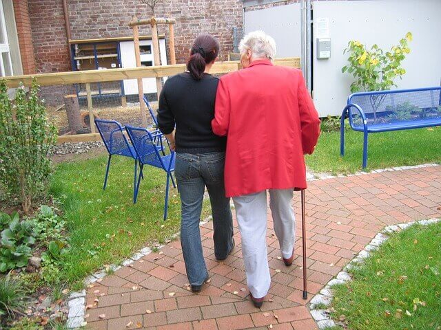 杖を持った高齢の女性が介護者と腕を組んで公園を散歩する写真